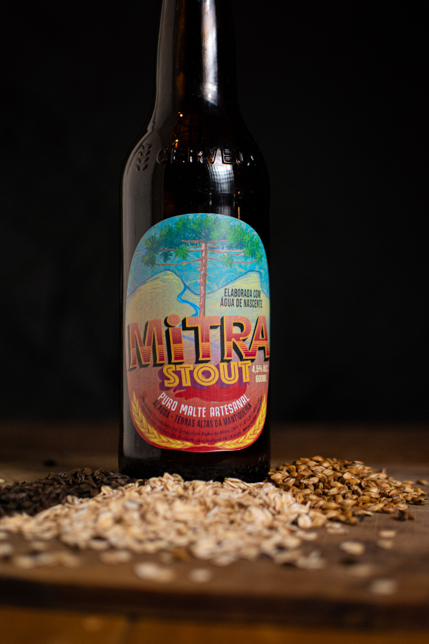Garrafa da Mitra Stout com rótulo vermelho sobre uma mesa com grãos de malte